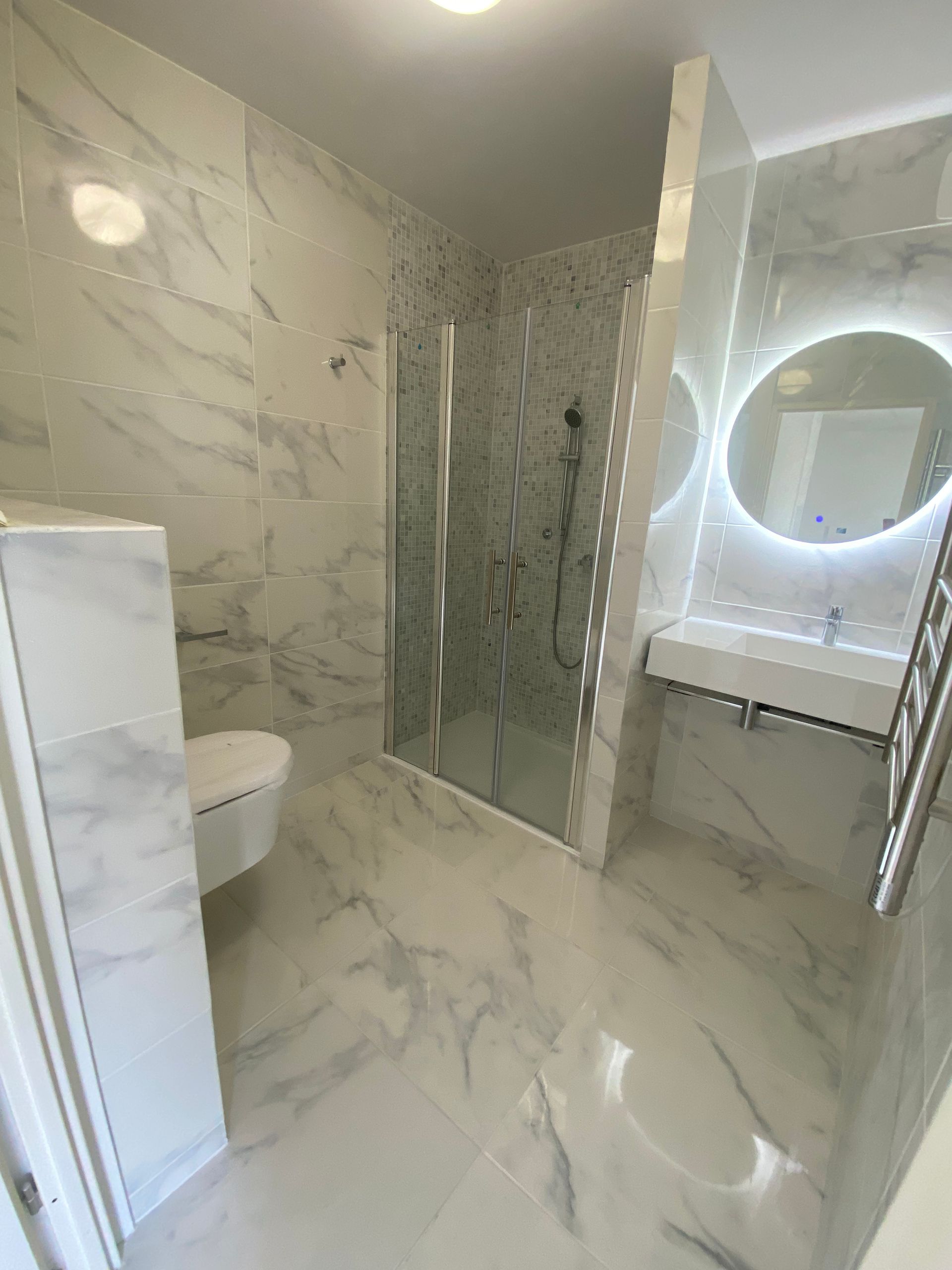 Salle de bain moderne avec carrelage aspect marbre, douche à l'italienne, lavabo flottant, miroir rond et toilettes.