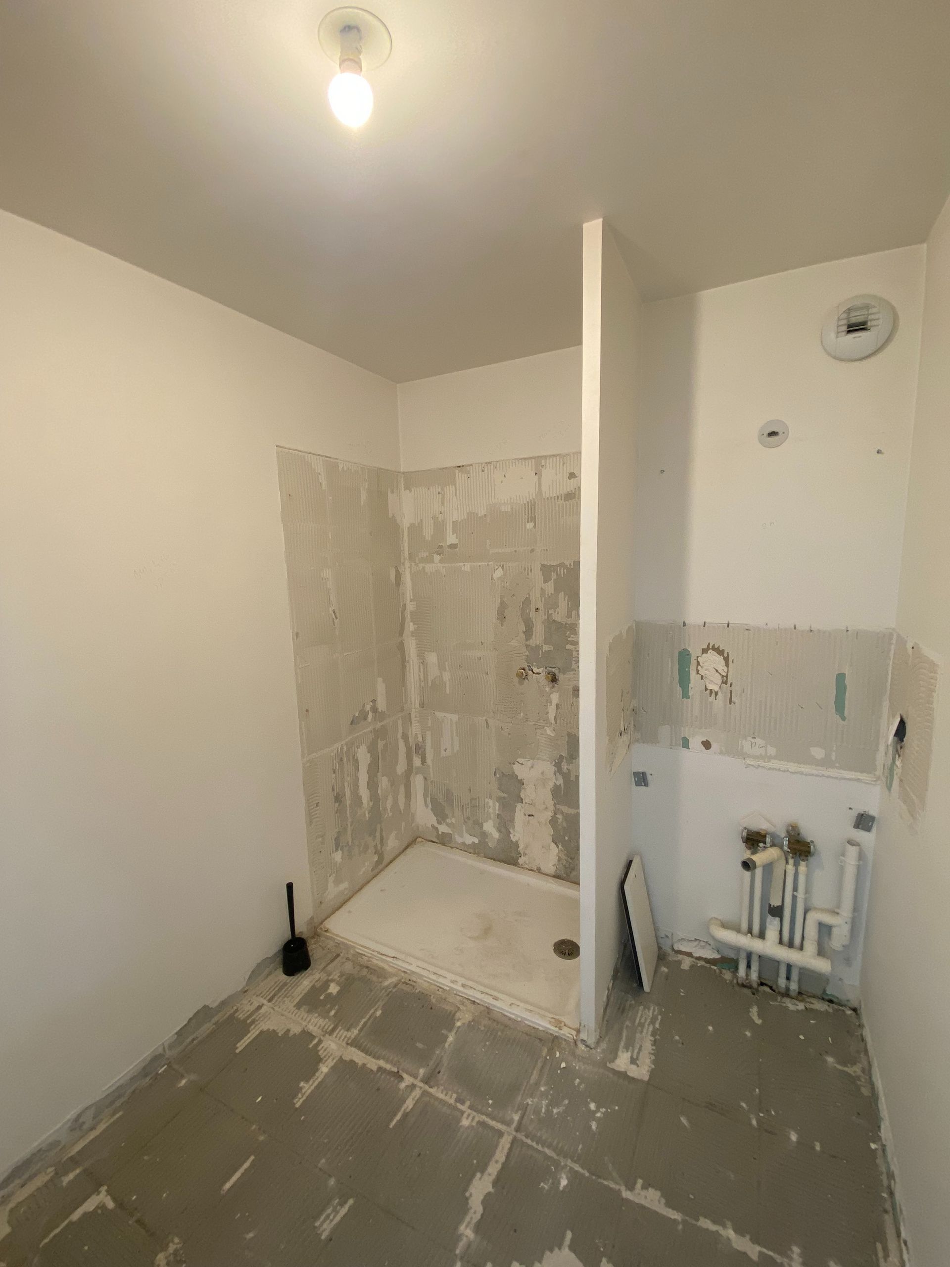 Rénovation de salle de bain en cours : cabine de douche, demi-mur, plomberie apparente. Murs et sol non finis, luminaire.
