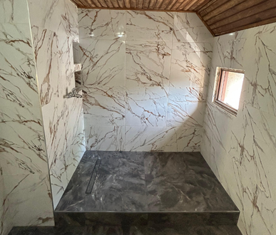 Douche carrelée en marbre avec sol gris foncé et petite fenêtre; plafond en bois.