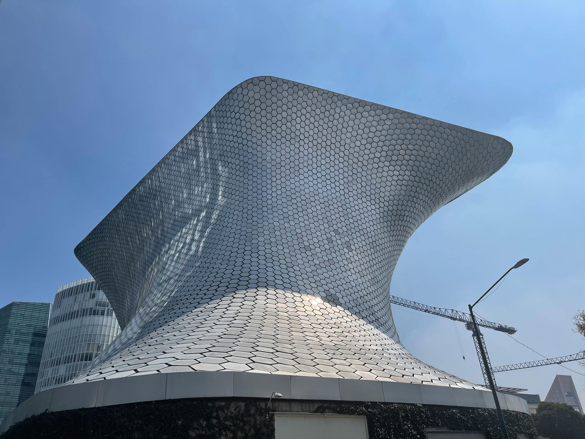Curved silver modern building with a textured facade against a blue sky, street crane nearby