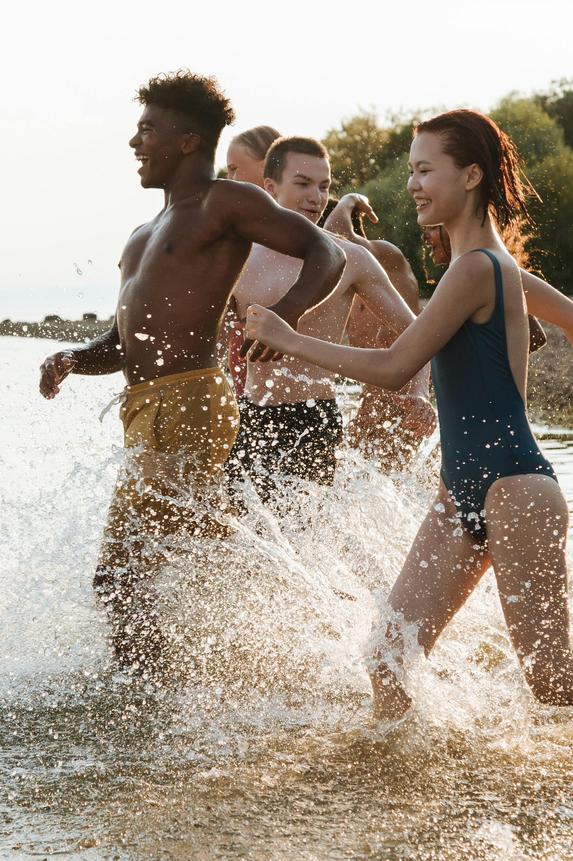 A group of friends laughing and splashing through shallow water at a beach during a sunset.