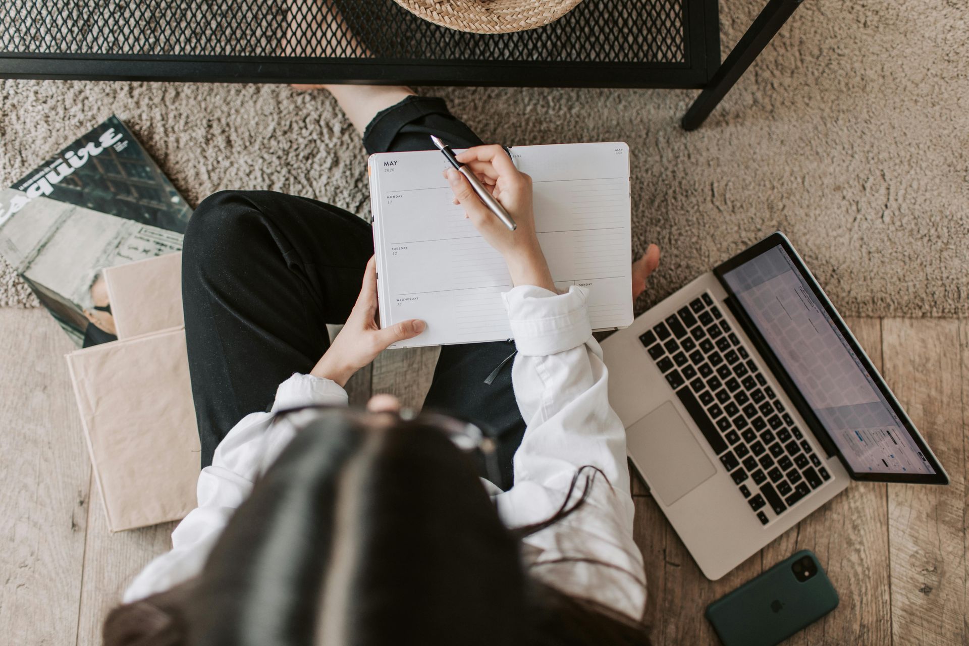 Person sitting on floor writing in a planner with laptop and phone nearby.