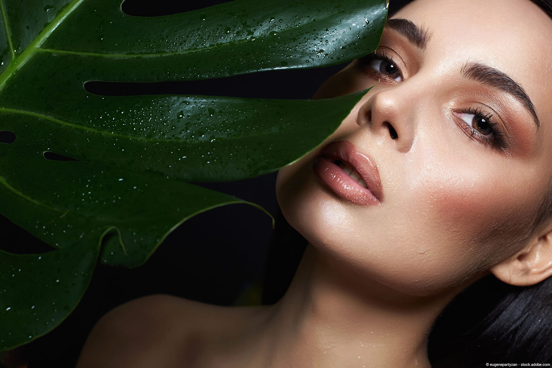 Gesicht einer Frau mit taufrischer Haut, teilweise verdeckt von einem großen grünen Blatt, mit bronzefarbenem Augen-Make-up.