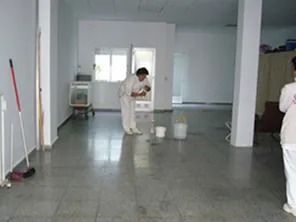 Persona fregando una habitación grande y vacía. Paredes blancas, suelo de baldosas grises.