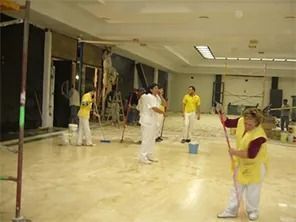 Gente limpiando una habitación grande con trapeadores y baldes, piso de mármol, algunos visten chalecos amarillos.