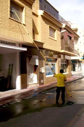 Un hombre con camisa amarilla lava las ventanas de un edificio con un palo largo y una manguera de agua en una calle mojada.