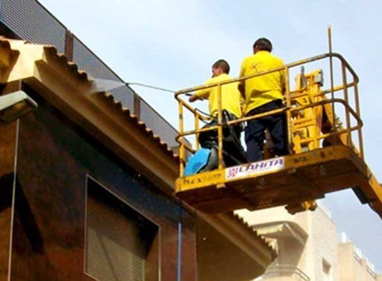 Dos trabajadores con camisetas amarillas lavan a presión el techo de un edificio desde un ascensor.