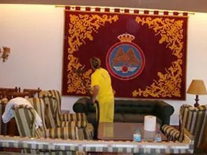 Una persona con una camisa amarilla se encuentra cerca de un sofá frente a un tapiz color burdeos con un escudo.