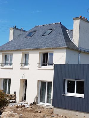 Maison à étage avec toiture en ardoise et extension grise ; chantier devant.