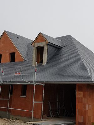 Une maison partiellement construite avec un toit gris foncé et des murs en briques orange ; échafaudage visible.