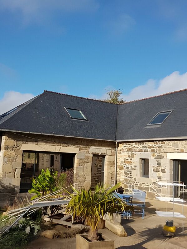 Maison en pierre avec un toit sombre et deux puits de lumière sur fond de ciel bleu, mobilier de jardin au premier plan.