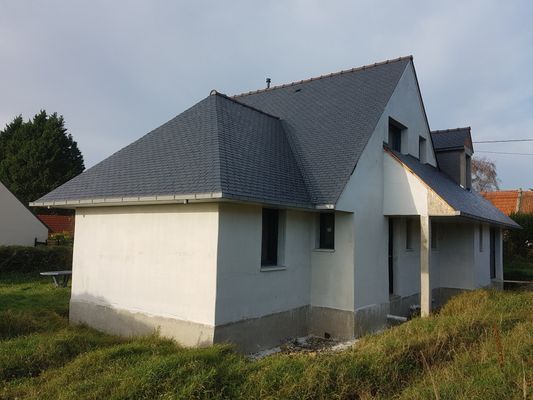 Une maison construite avec un toit en ardoise grise et des murs extérieurs blancs, située dans une cour herbeuse.
