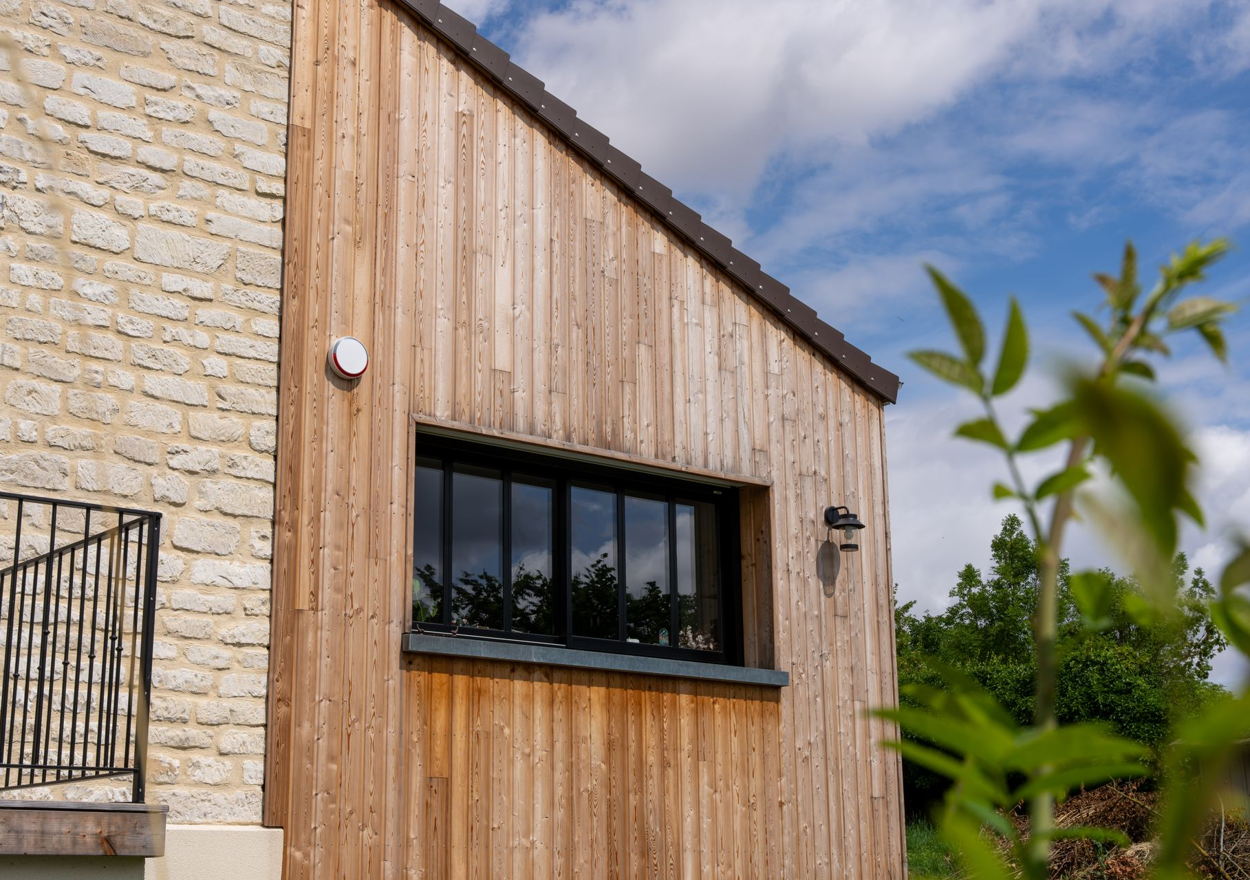 Vue extérieure d'un bâtiment avec bardage en bois, fenêtre noire et mur en pierre. Ciel nuageux.