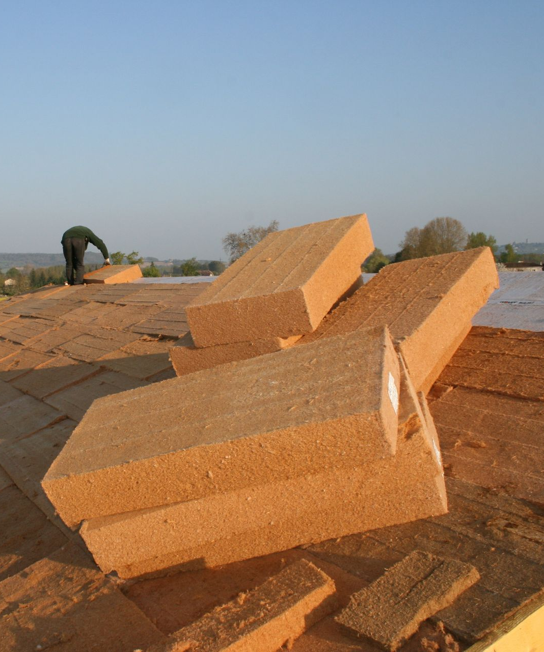 Un ouvrier du bâtiment installe des panneaux isolants sur un toit, avec des planches empilées au premier plan.
