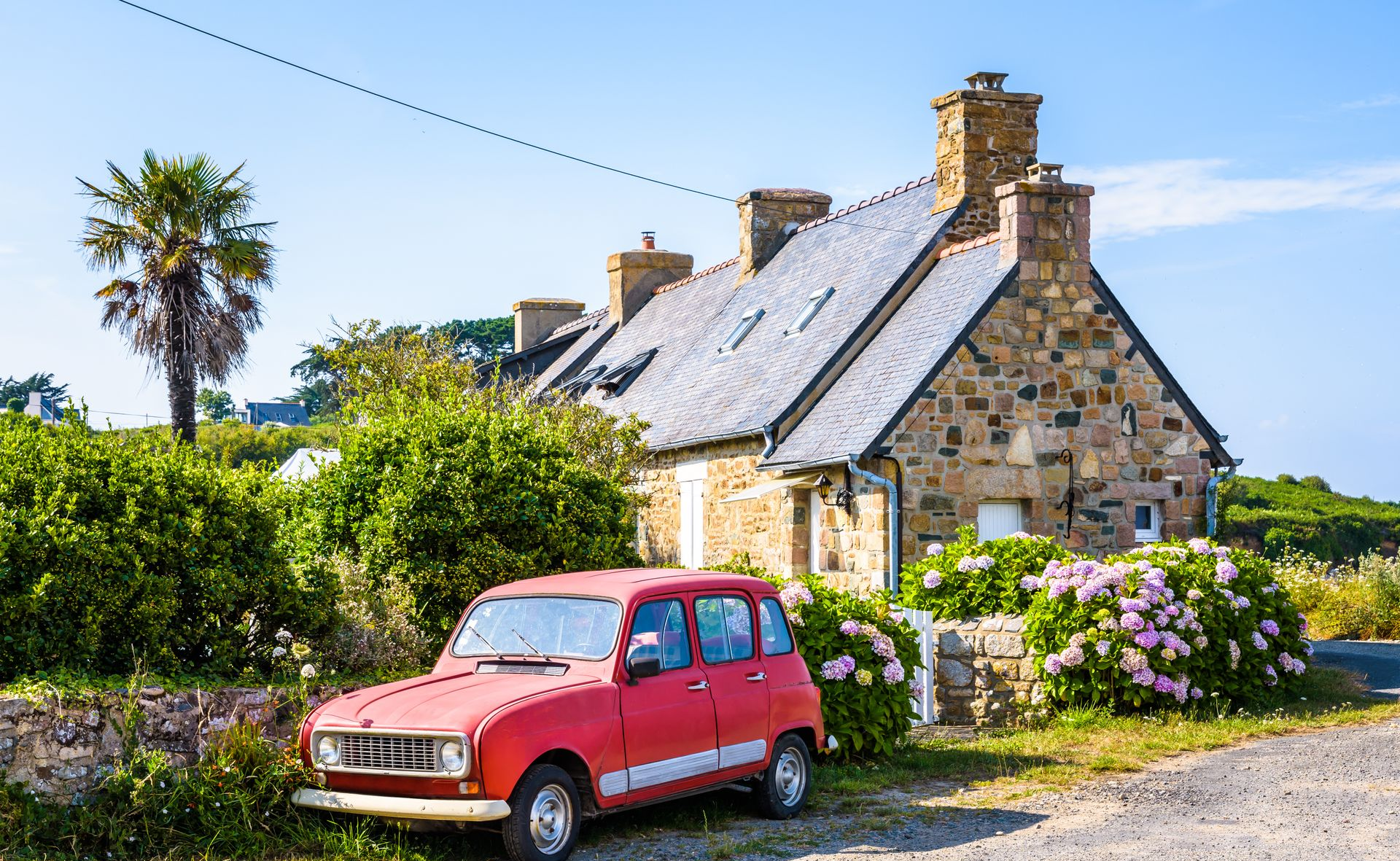 Voiture rouge garée devant un chalet en pierre avec une cheminée et des buissons par une journée ensoleillée.