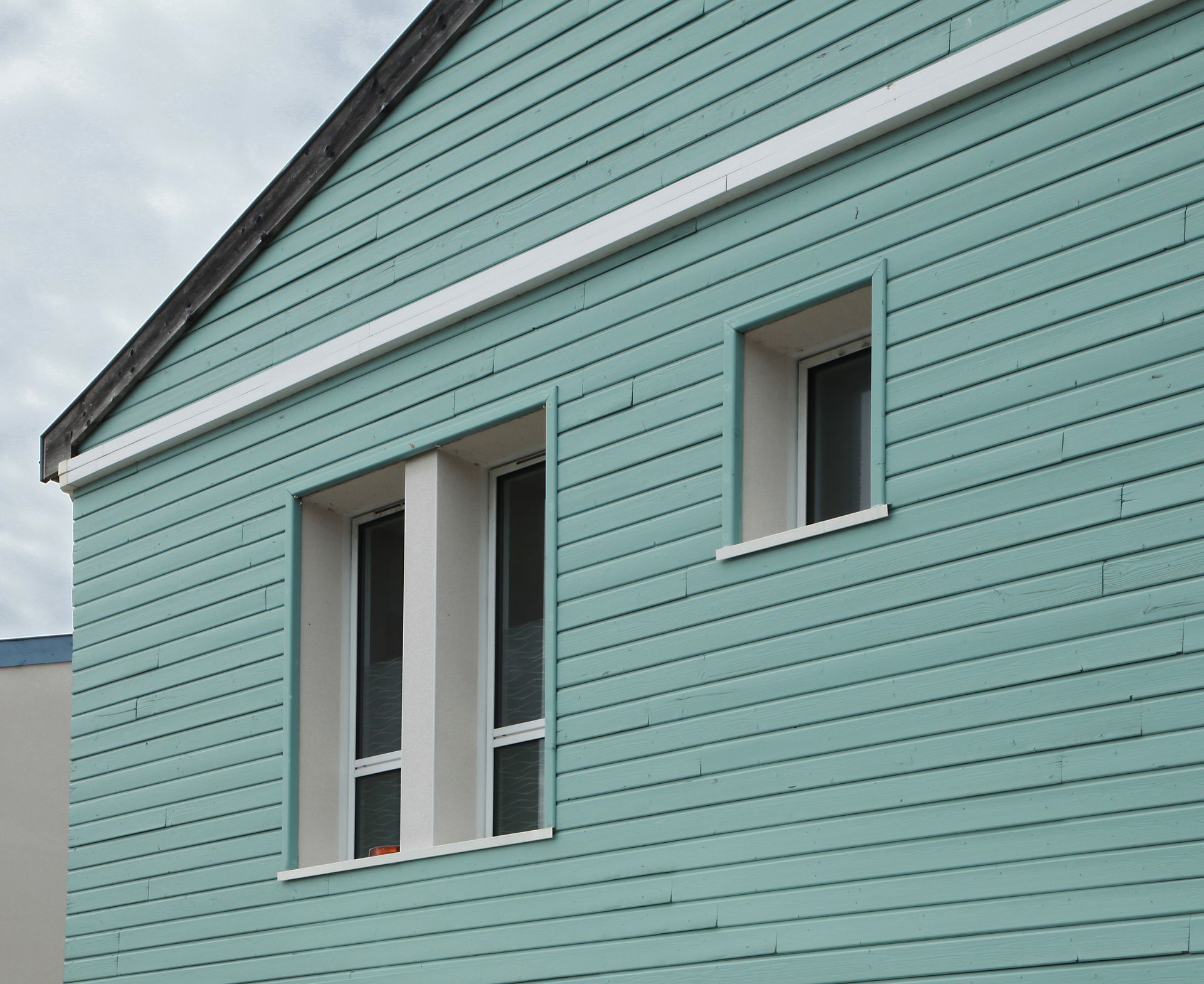 Bâtiment de couleur sarcelle avec des fenêtres à cadre blanc, contre un ciel nuageux.