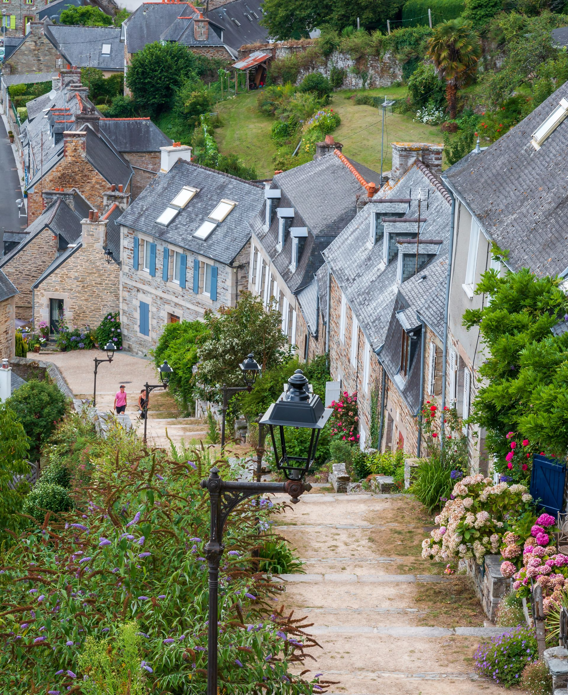 Des bâtiments en pierre bordent une rue pavée, un écrin de verdure et un lampadaire. Deux personnes marchent.