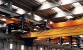 Interior de una nave industrial con vigas de grúa aéreas de color naranja y cerchas metálicas en el techo.