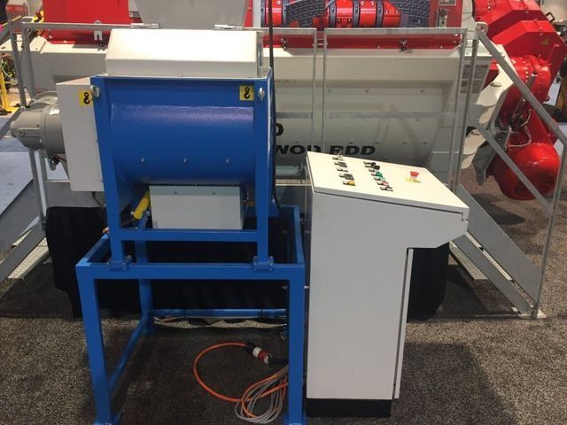 Máquina industrial azul junto a un armario de control blanco en una plataforma de fábrica con tuberías rojas.