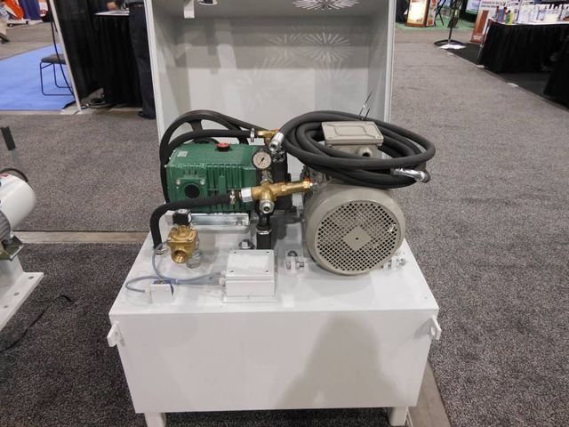 Pequeño compresor de aire verde con manguera enrollada y motor sobre un pedestal blanco en un stand de exposición.