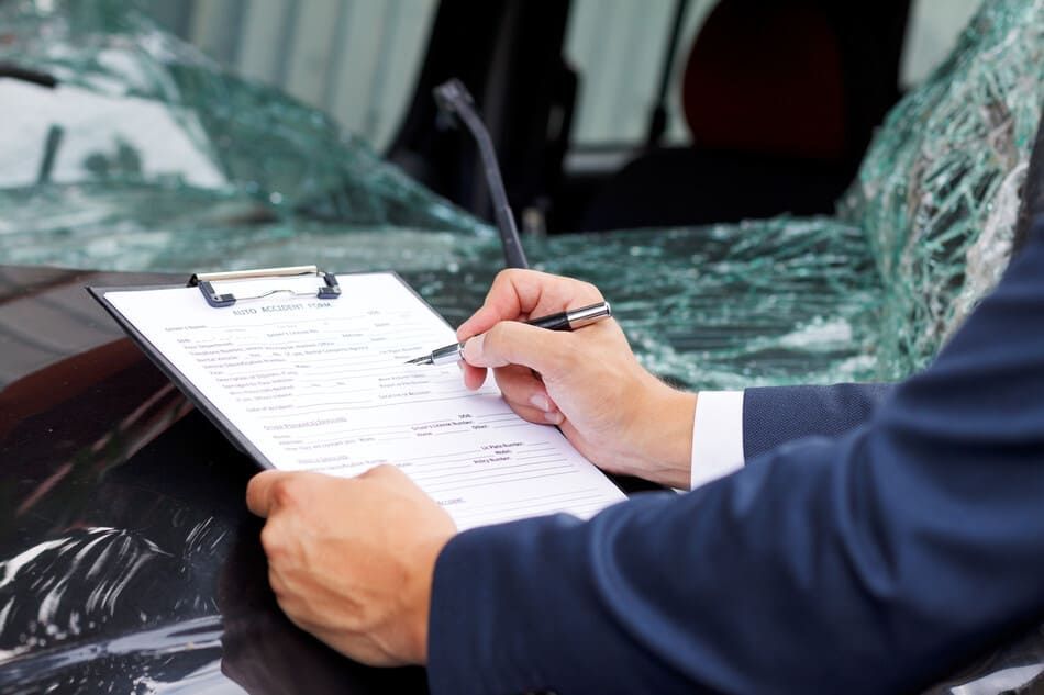 Un hombre está escribiendo en un portapapeles delante de un coche con el parabrisas roto.