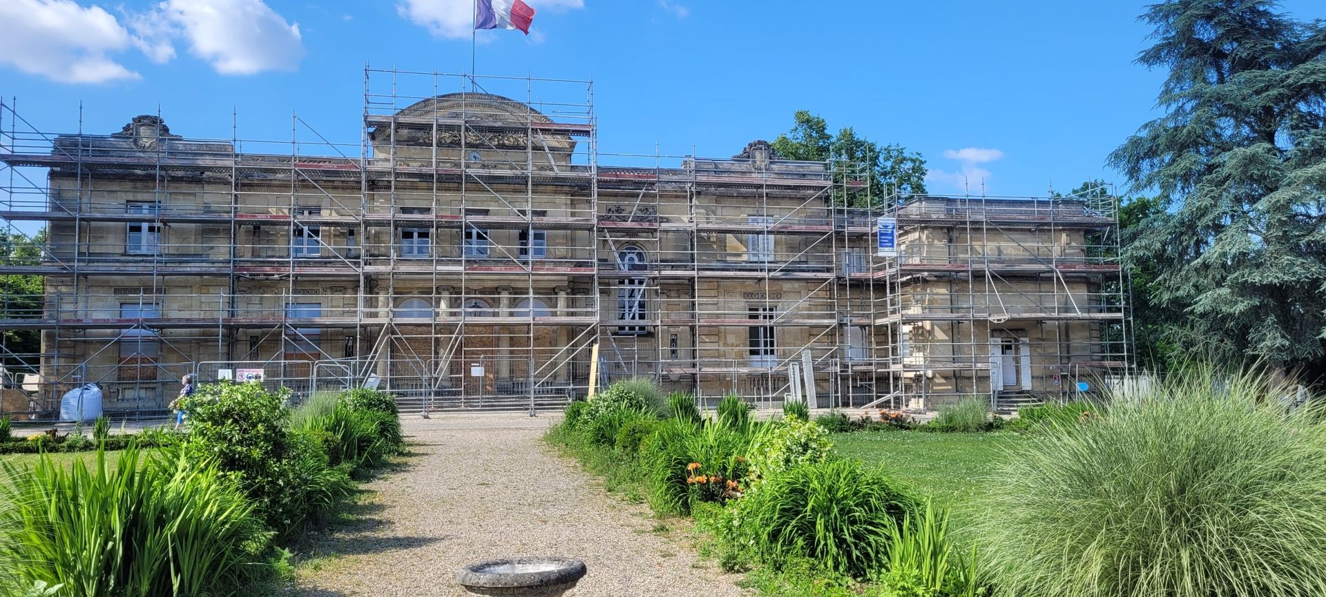 Un grand bâtiment en pierre en rénovation, avec des échafaudages, un chemin et de la végétation.
