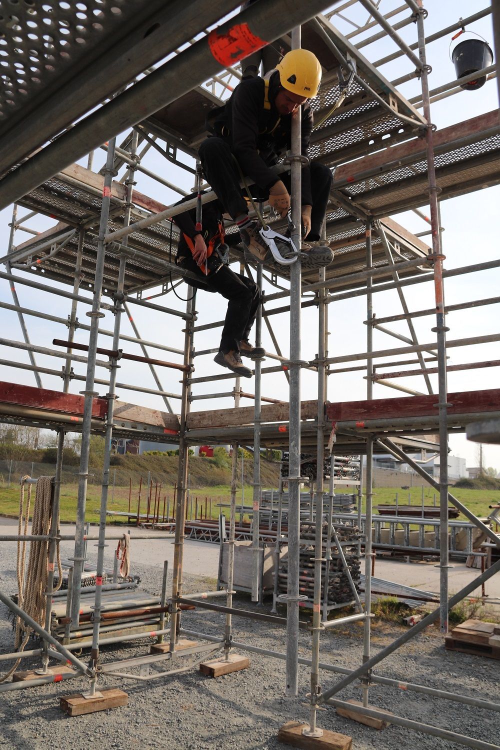Personne sur un échafaudage, portant un harnais et un casque, en train de sécuriser du matériel.