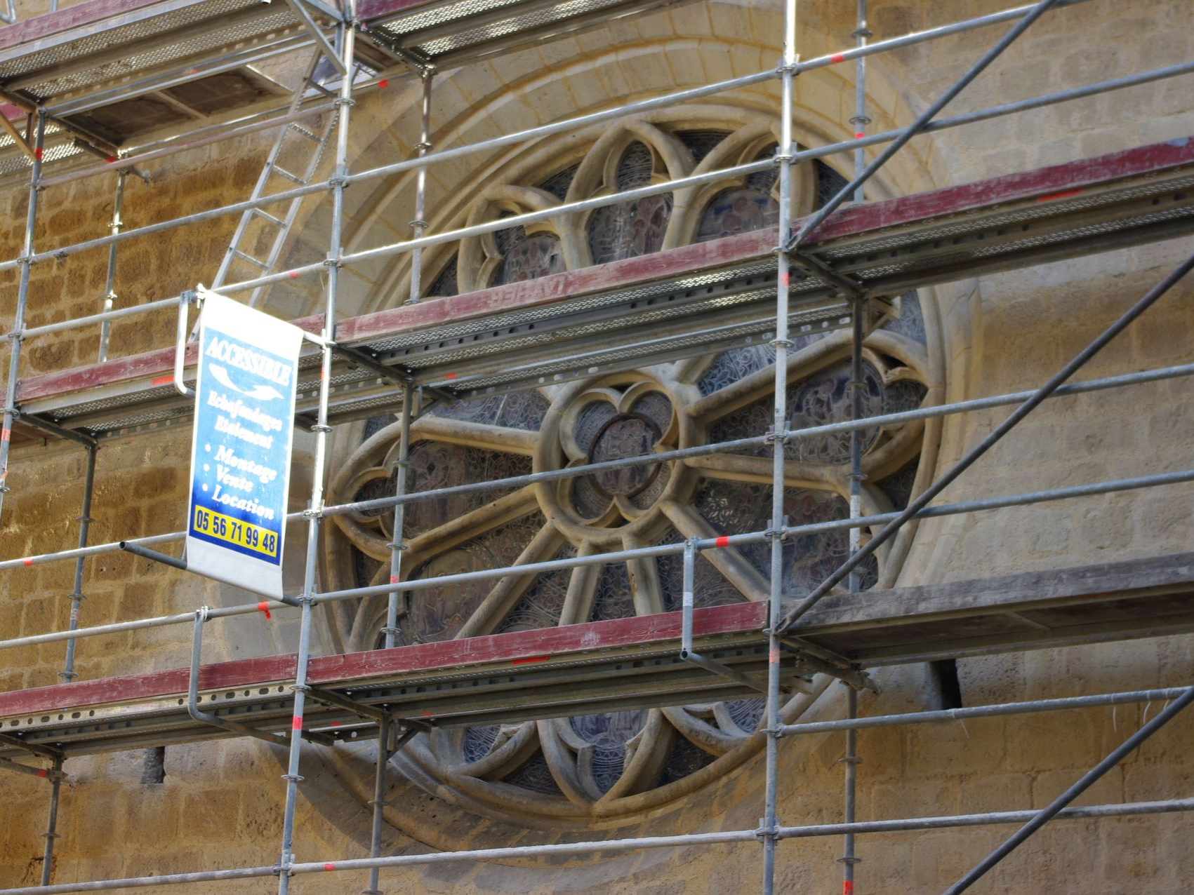 Des échafaudages entourent un vitrail rond dans un bâtiment en pierre. Une pancarte est accrochée à l'échafaudage.