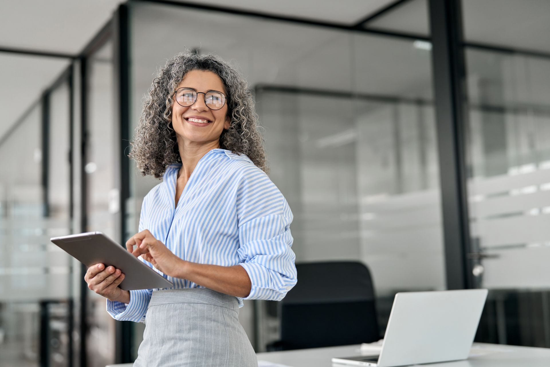 Femme à lunettes, tablette à la main, souriante au bureau. Chemise bleu clair à rayures, jupe grise.