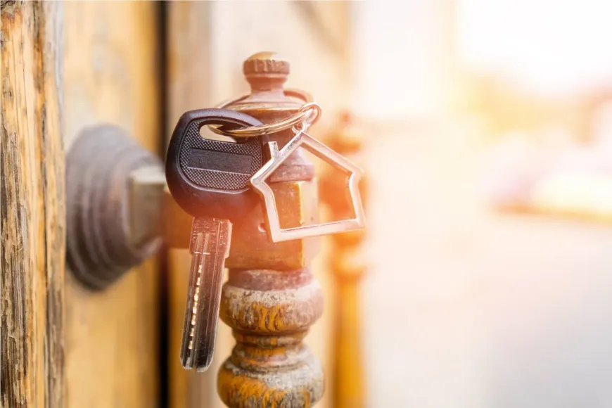 Llaves en una manija de puerta antigua con un llavero con forma de casa, luz solar brillante en el fondo.