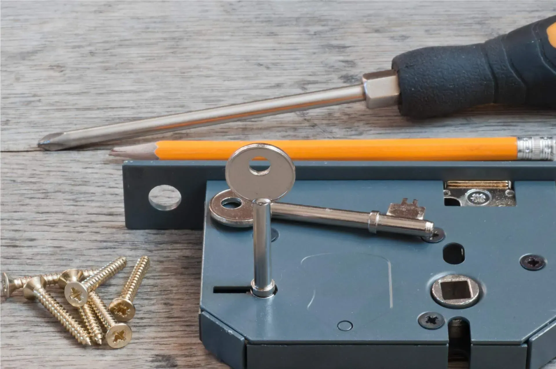 Cerradura con llave y herramientas: destornillador, lápiz y tornillos. Cerradura gris sobre una superficie de madera.