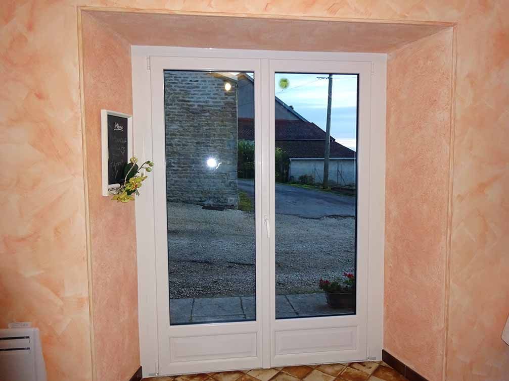 Des portes doubles vitrées blanches dans une pièce couleur pêche donnent sur une allée et une maison.