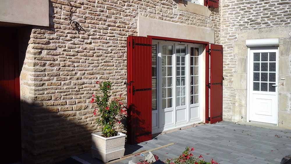 Maison en pierre avec volets rouges et porte-fenêtre blanche donnant sur une terrasse