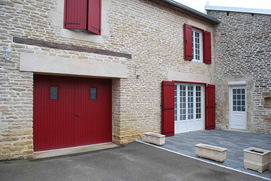 Façade en pierre d'une maison avec garage et volets rouges, fenêtre à cadre blanc et cour pavée.