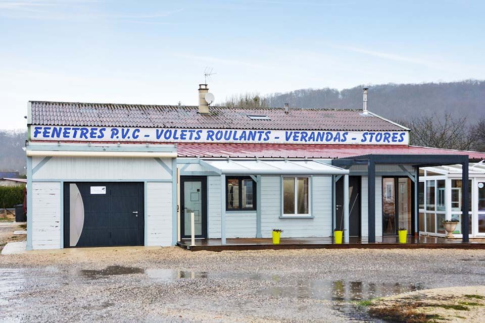 Façade d'un commerce de portes et fenêtres avec une enseigne bleue et un extérieur gris sous un ciel couvert