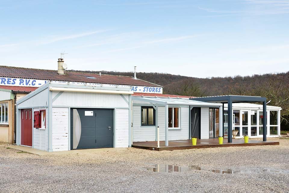 Petit bâtiment blanc avec une porte de garage grise, des fenêtres et une véranda vitrée, sur un terrain gravillonné sous un ciel dégagé.