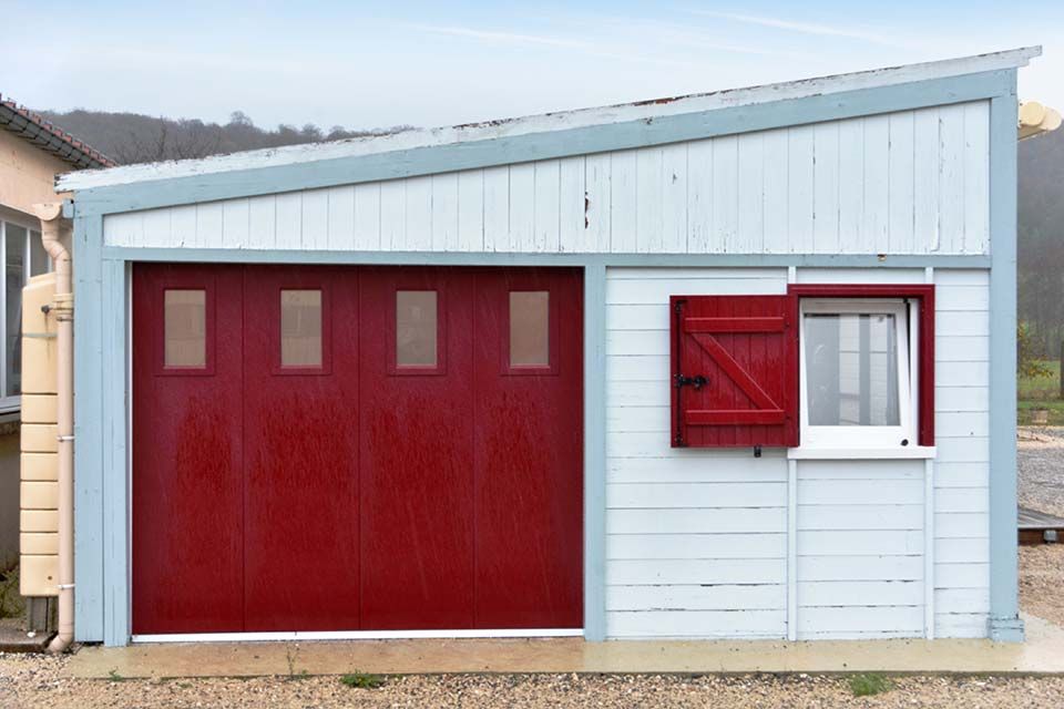 Abri de jardin bleu clair avec porte de garage rouge et fenêtre à encadrement rouge sur un terrain en gravier
