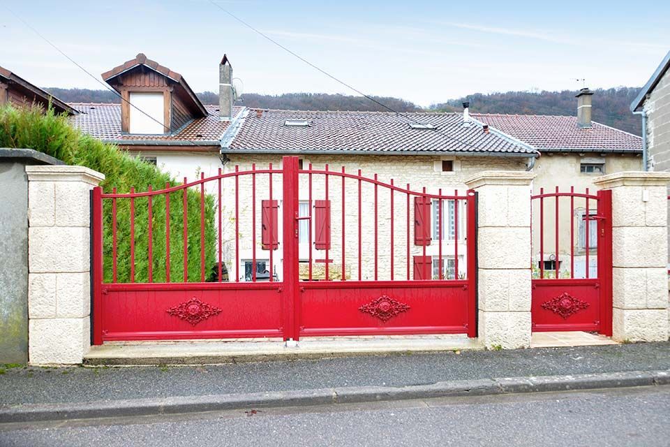 Portail d'allée en métal rouge devant une maison en pierre au toit de tuiles