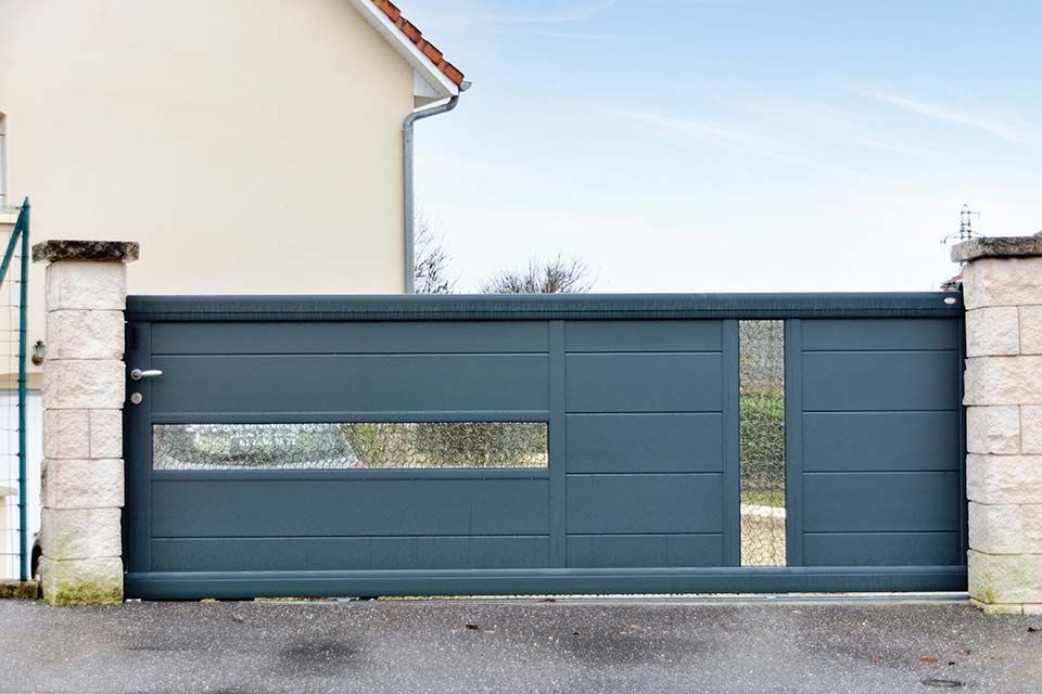 Portail coulissant bleu-gris avec une fenêtre étroite, placé entre des piliers en pierre à côté d'une maison
