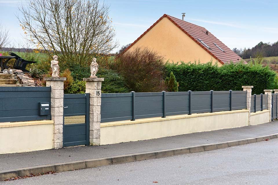 Maison de banlieue derrière un mur couleur crème et une clôture sombre, avec un toit de tuiles et des arbres le long d'une rue calme.