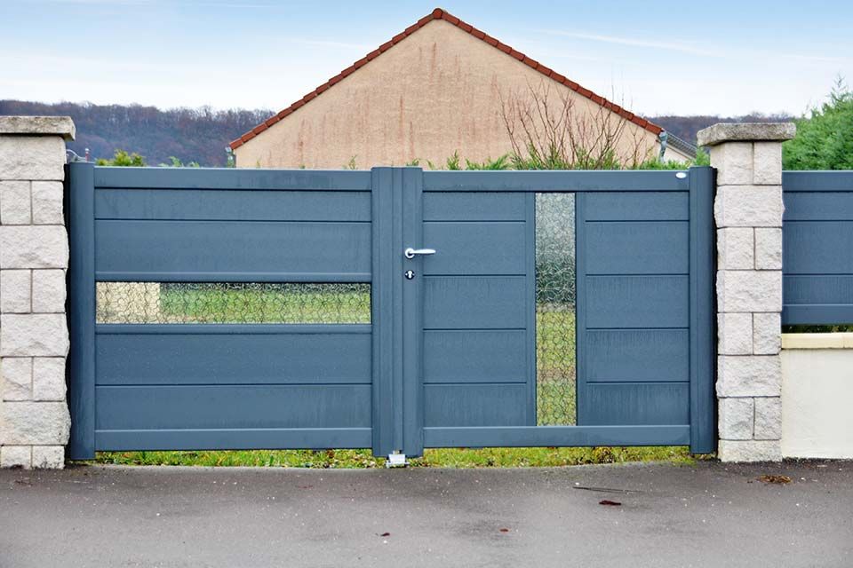 Portail en métal bleu avec une porte centrale entre deux piliers de pierre, devant une maison et une cour gazonnée