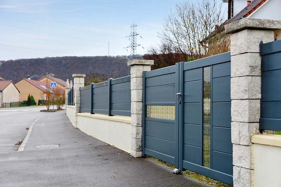 Rue résidentielle bordée d'un long mur gris et d'un portail, donnant sur une route mouillée, avec des maisons en arrière-plan.