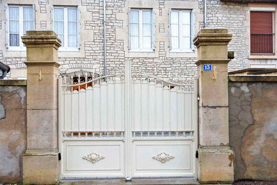 Un portail en fer forgé blanc entre des piliers de pierre devant un bâtiment en briques claires