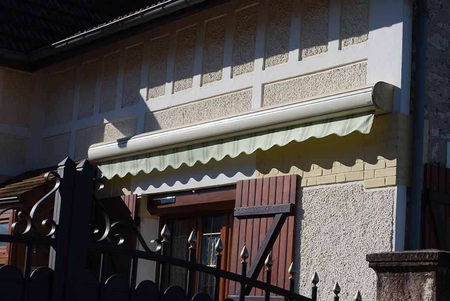 Façade de maison avec un auvent festonné au-dessus d'une porte et d'une fenêtre en bois, derrière un portail en métal noir