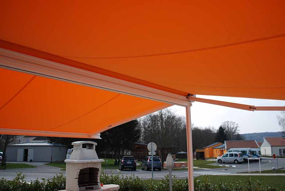 Un auvent orange recouvre une terrasse, avec un chauffage blanc et des voitures garées sur un parking en bord de route.