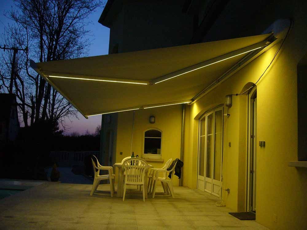 Patio éclairé de jaune avec un auvent blanc, une table et des chaises, à l'extérieur d'une maison au crépuscule