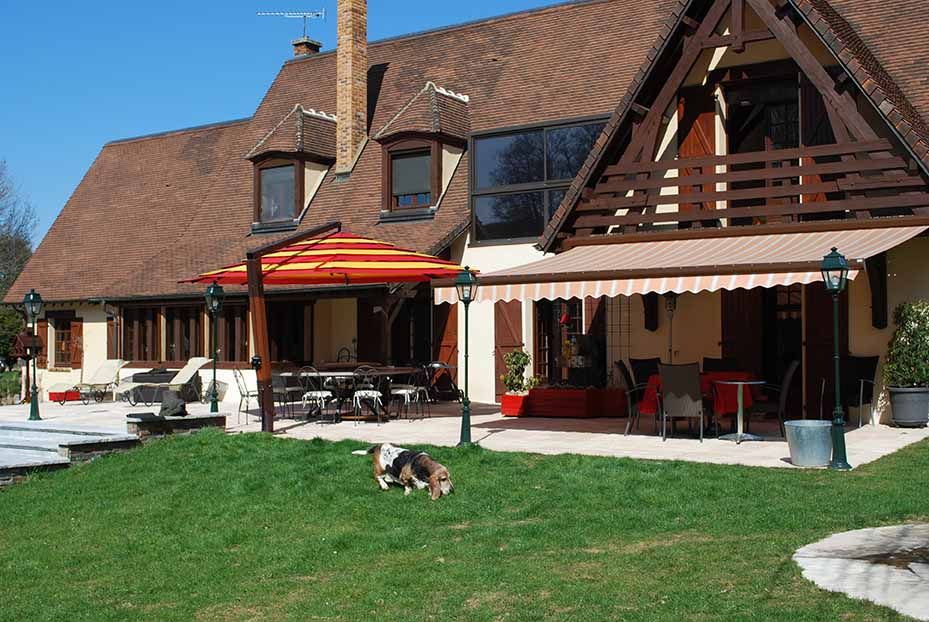 Maison en briques avec parasols de terrasse, chaises rouges et un chien sur la pelouse verte.
