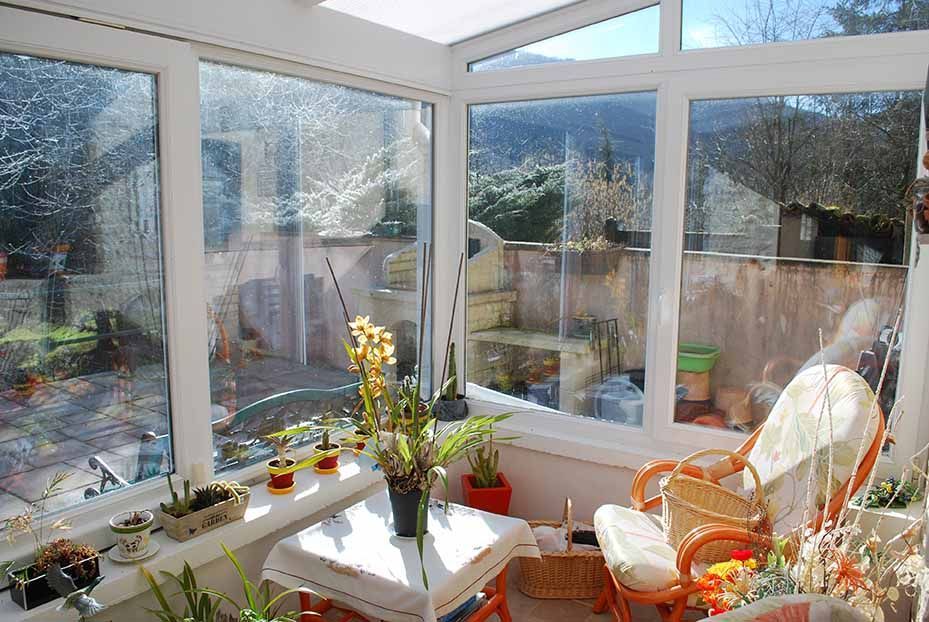 Véranda baignée de soleil avec fauteuils en osier, plantes en pot et petite table donnant sur un jardin