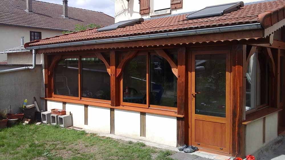 Véranda en bois avec de grandes fenêtres cintrées et une porte vitrée, attenante à une maison, à côté d'une petite pelouse.