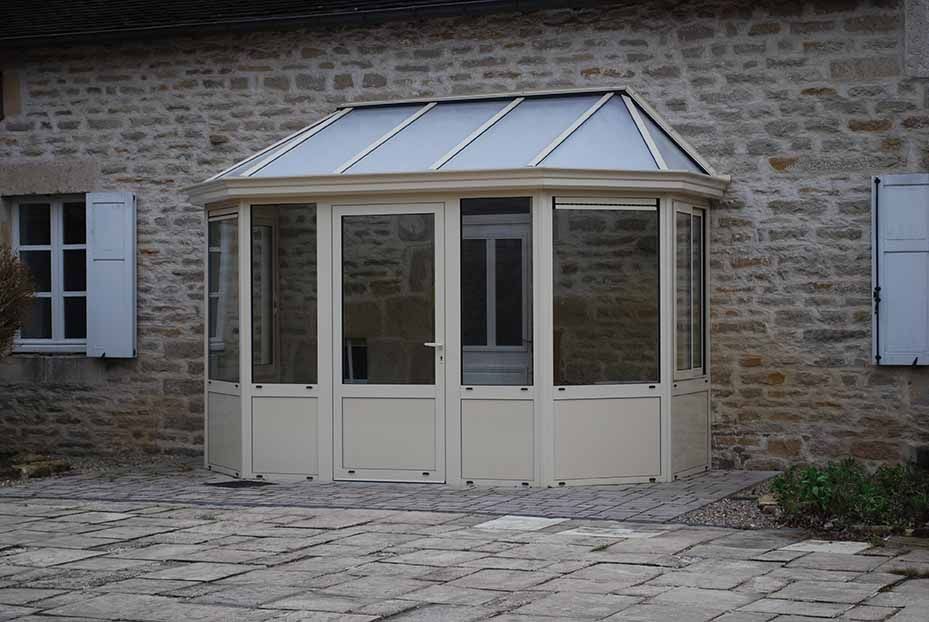 Véranda à toit de verre attenante à un bâtiment en pierre, avec fenêtres et portes à cadre blanc donnant sur une cour pavée.